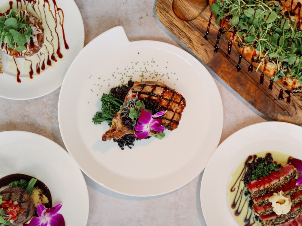 An overhead view of five elegantly plated gourmet dishes, including grilled meat, seared tuna, a small filet, a flatbread with greens, and an appetizer drizzled with sauce.