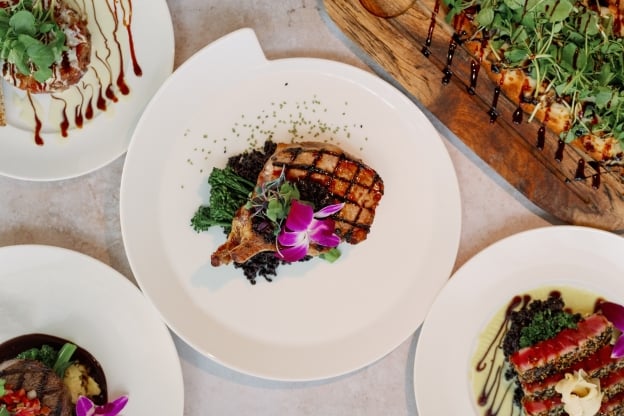 An overhead view of five elegantly plated gourmet dishes, including grilled meat, seared tuna, a small filet, a flatbread with greens, and an appetizer drizzled with sauce.